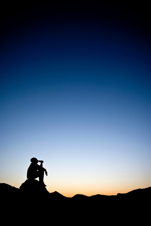 A male hiker silhouetted by a dawn sky views a mountain landscape as he drinks from a water bottle while seated on a rock.