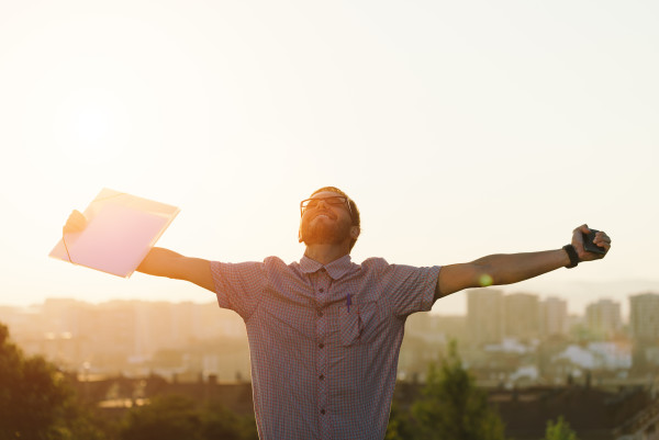 Successful professional casual man celebrating work success and raising arms against city background on sunset. Job achievement or goal concept.