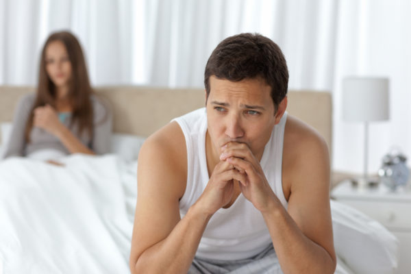 Pensive man having a headache sitting on the bed with his girlfriend on the background